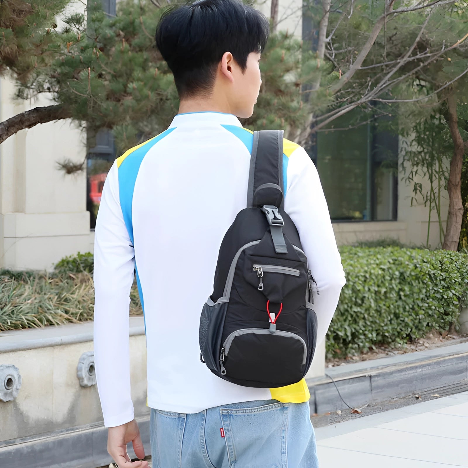 Crossbody sling backpack in a vibrant yellow color, featuring a water-resistant material and a USB port, worn over the shoulder by a person wearing jeans. The lightweight bag is set against a backdrop of greenery.