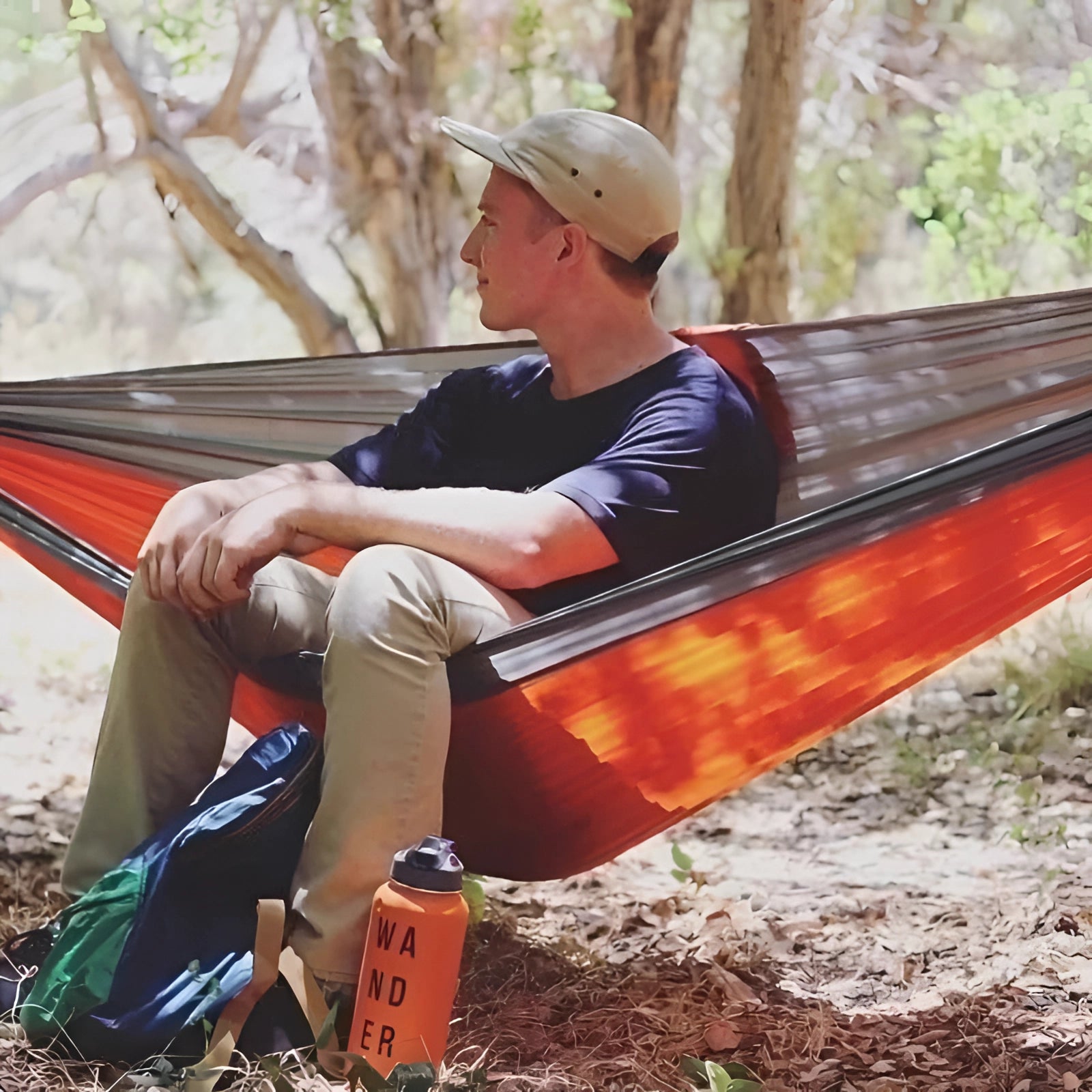 A double-layer insulated camping hammock in olive green and teal color, set up between trees in a natural outdoor setting with people nearby, featuring attached straps for support.