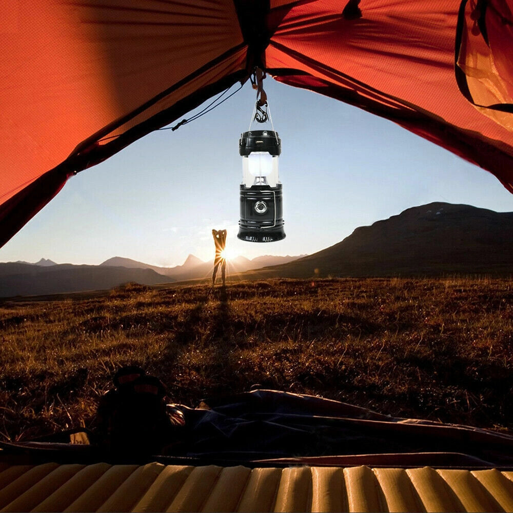 Dual-Function LED Camping Lantern & Rechargeable Flashlight - Waterproof illuminating a campsite next to a tent with mountains and a wooden area in the background under a clear sky.