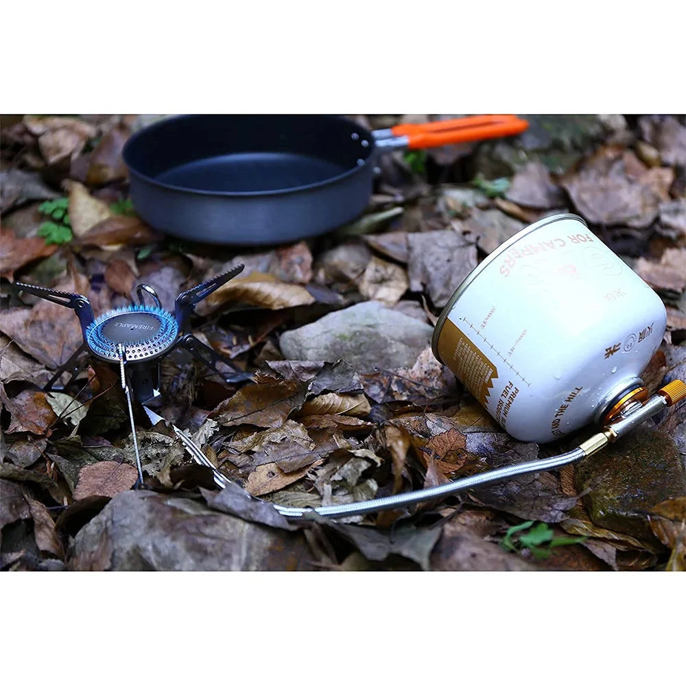 
Compact Fire-Maple Blade 2 camping stove set up on a wooden surface, featuring a metal windproof design with gas canister attached, surrounded by camping tableware and drinkware.