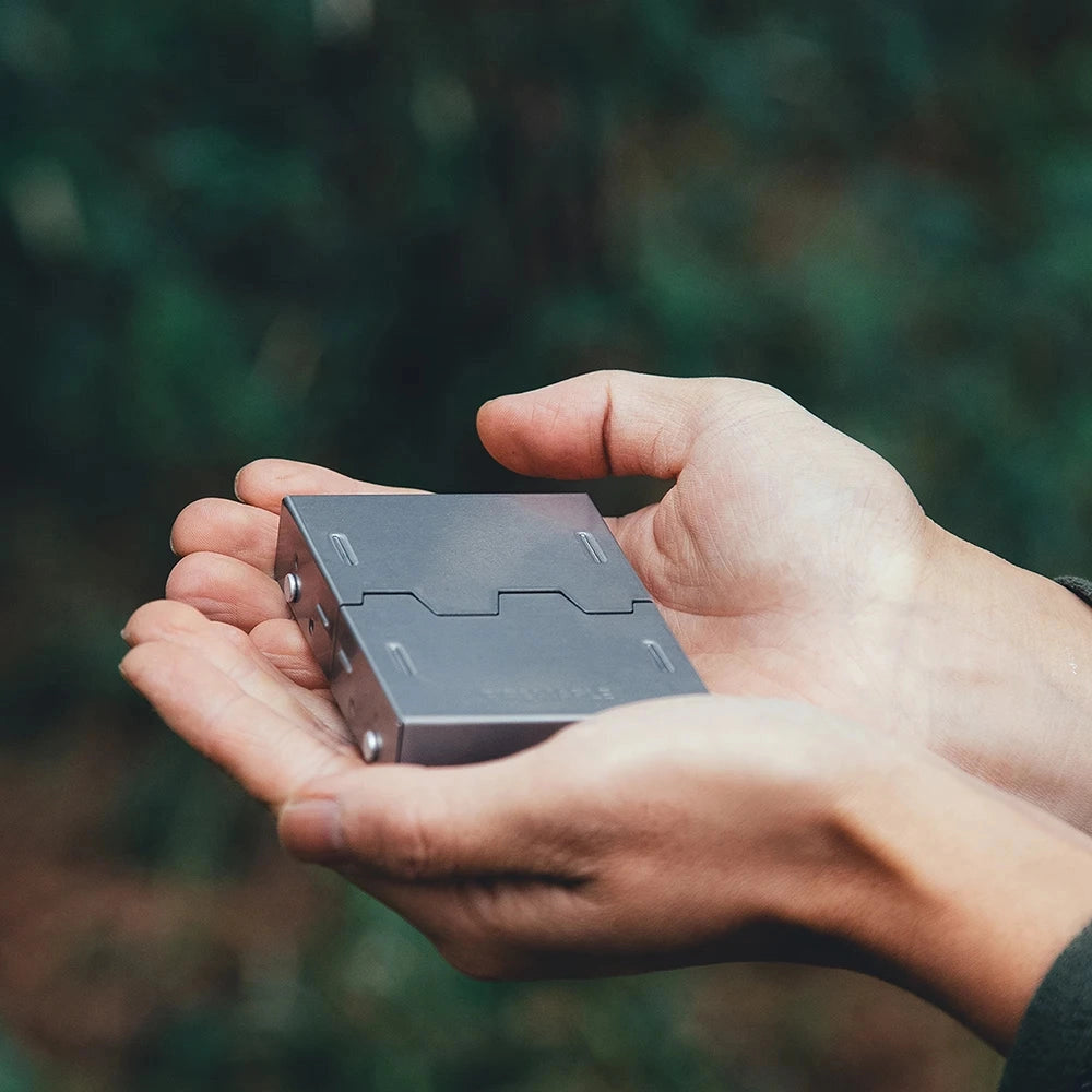 A person's hand holding the FireMaple Lotus Titanium Ultralight Alcohol Stove, designed for camping and hiking, with a focus on the compact and lightweight design. The hand is set against a wooden background, highlighting the stove's portability and ease of use.