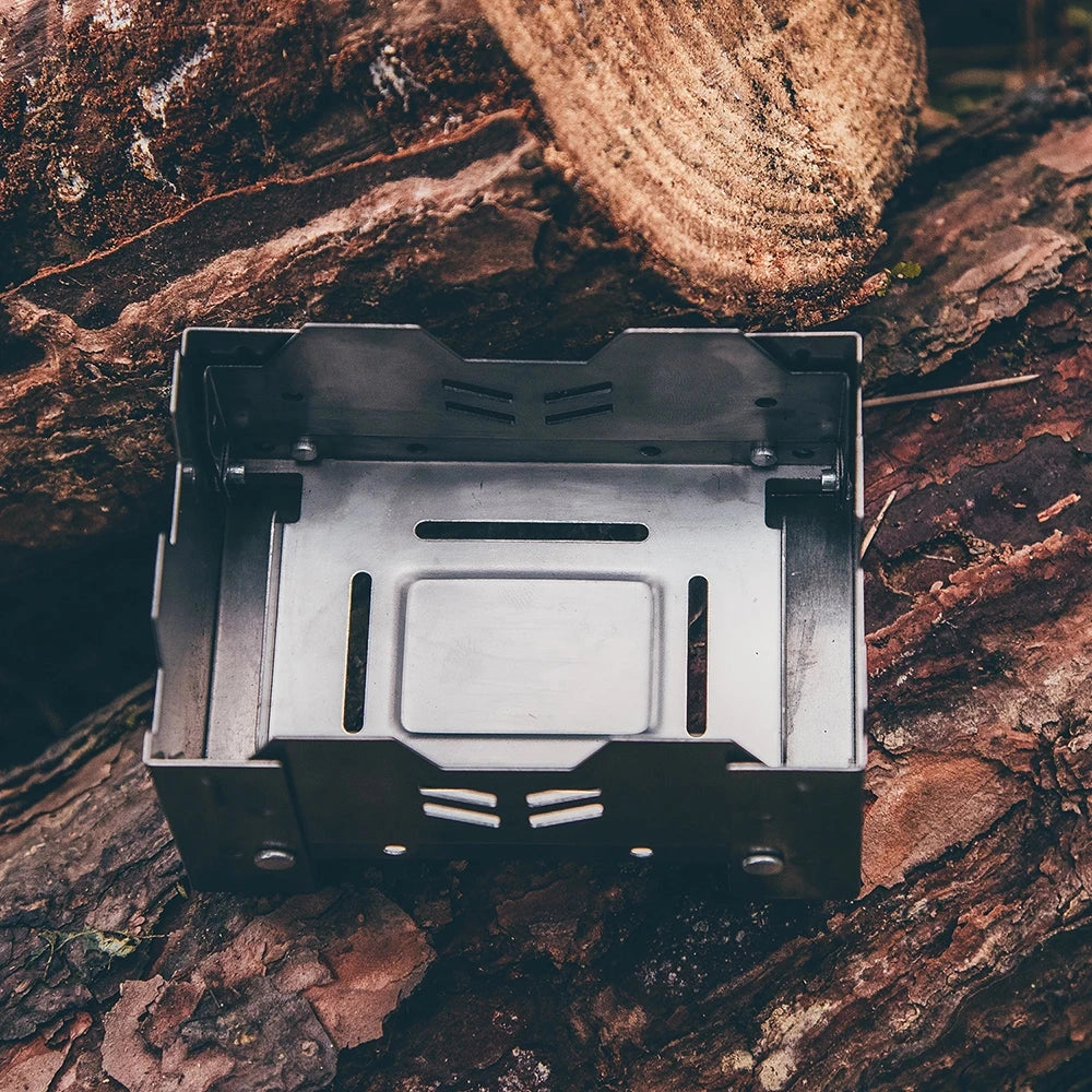 FireMaple Lotus Titanium Ultralight Alcohol Stove set up on a flat surface in an outdoor camping or hiking setting, surrounded by natural elements like trees and wood, with a focus on its compact and lightweight design suitable for outdoor cooking.