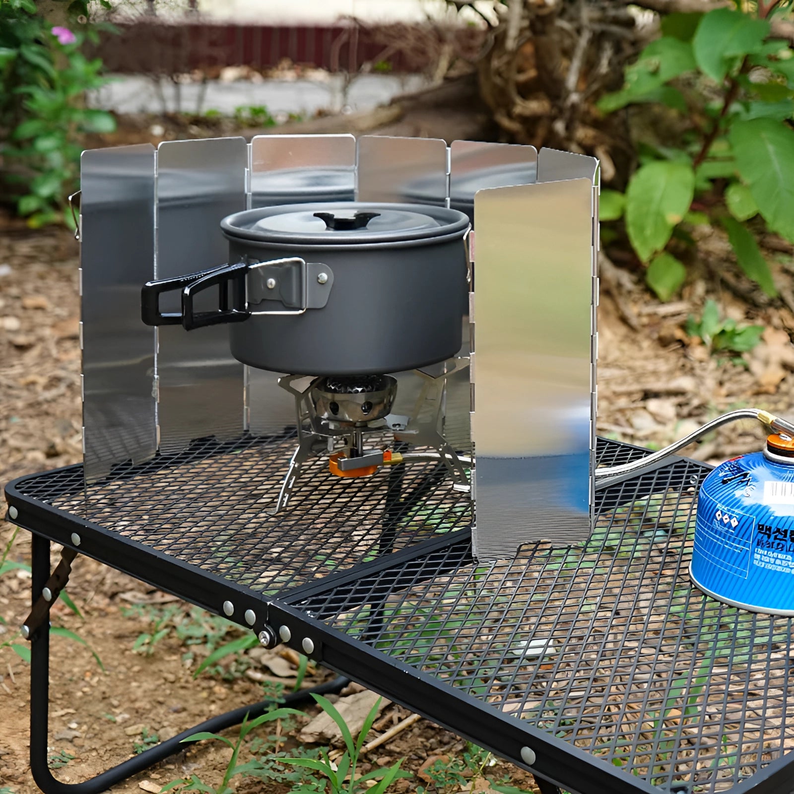 Foldable Aluminum Camping Stove Windscreen set up outdoors with a portable gas stove, surrounded by plants.