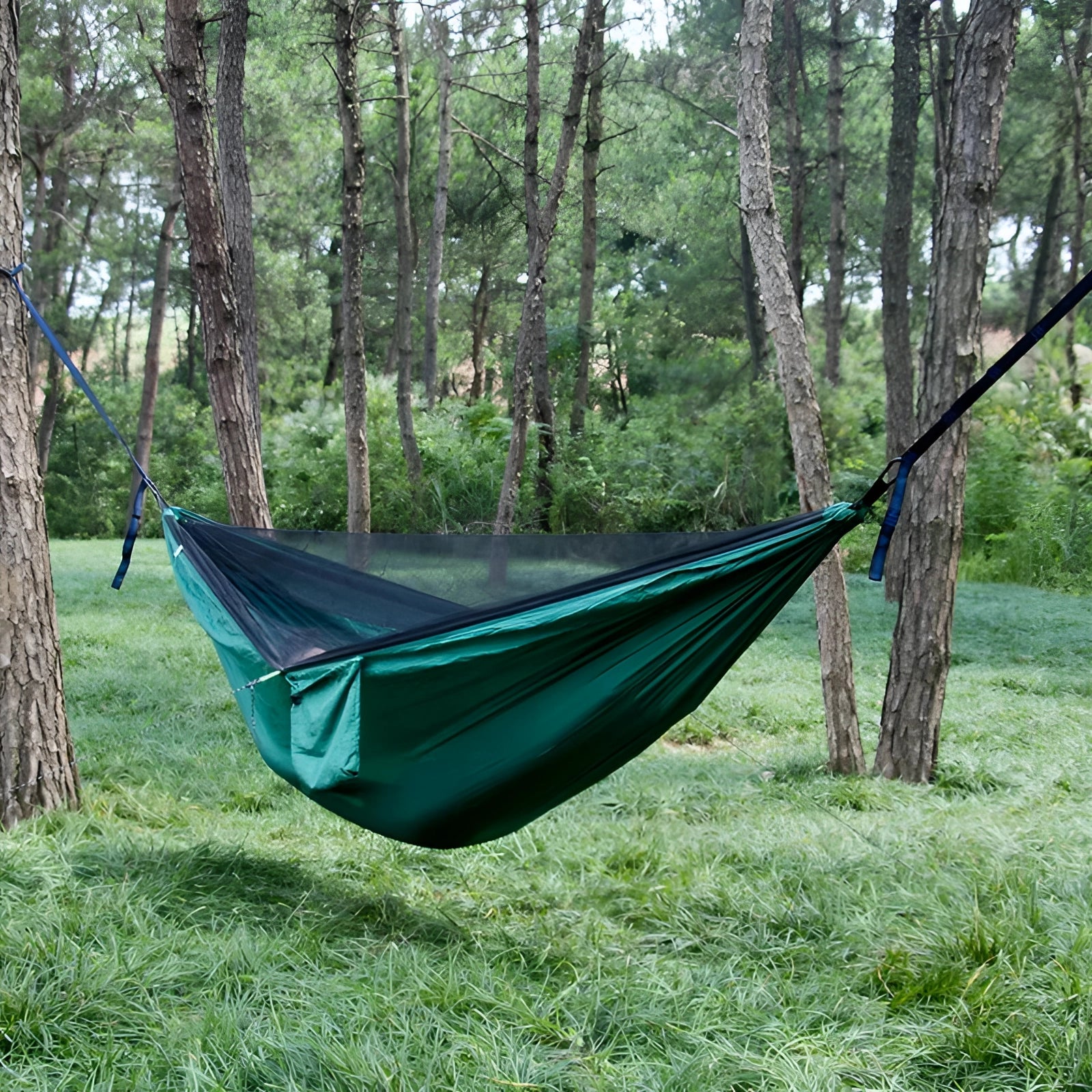 Green lightweight mosquito net hammock suspended between trees in a grassy area, set up for camping with tarpaulin providing shade in the background.