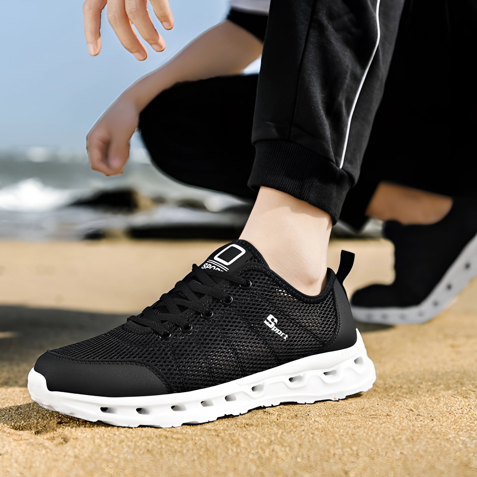 A pair of lightweight breathable mesh running shoes in black and white, shown on a person's feet, with visible hand, leg, and knee.