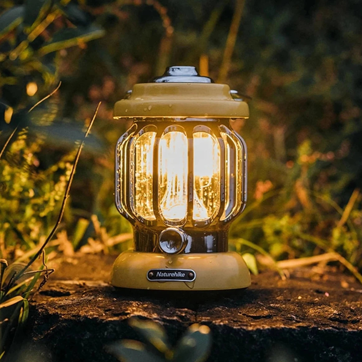 Vintage-style Naturehike LED camping lantern, portable and rechargeable, placed on the grass near trees and plants, illuminating the outdoor landscape.