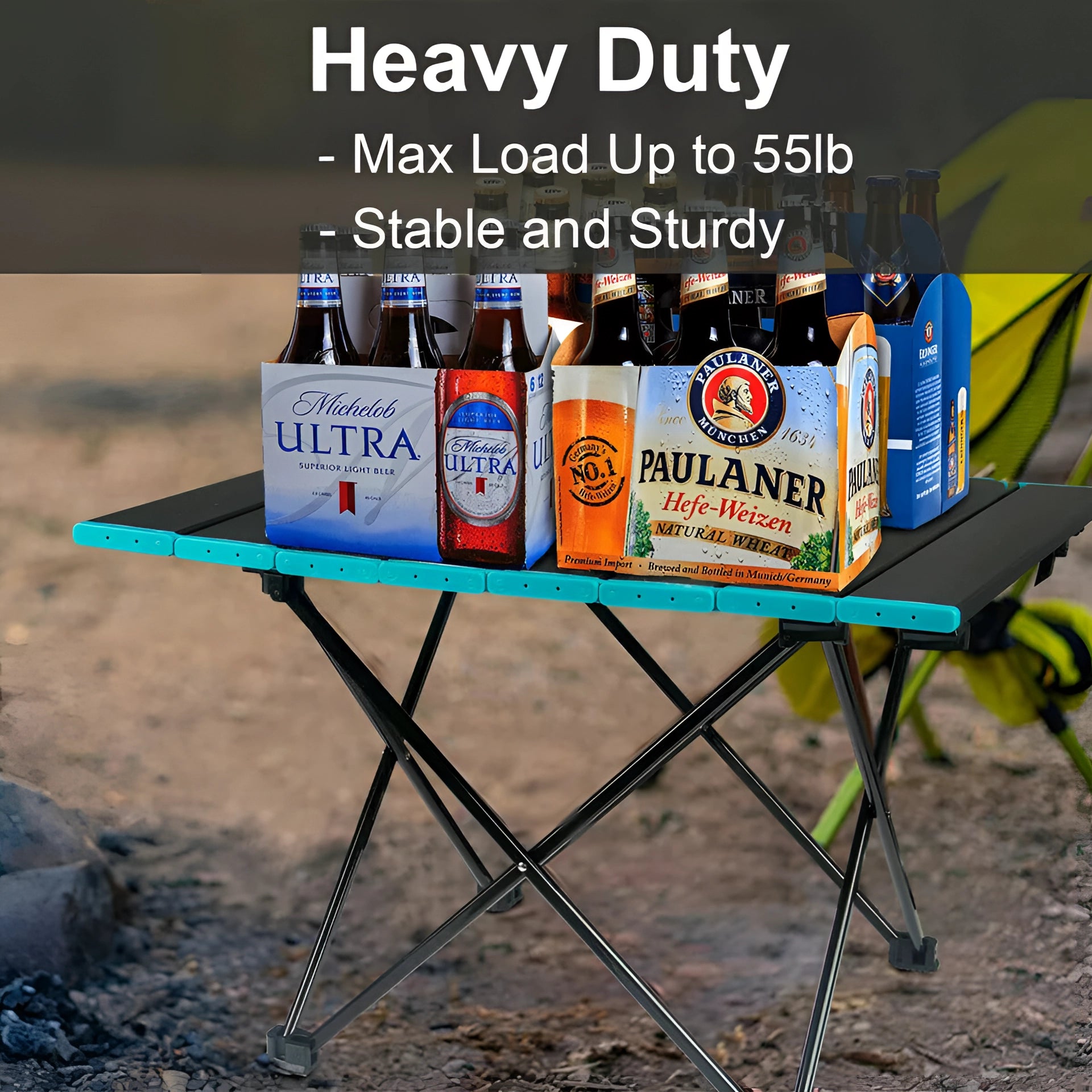 Portable black aluminum folding camping table with carrying bag set up outdoors with a bottle on top, surrounded by natural landscape.