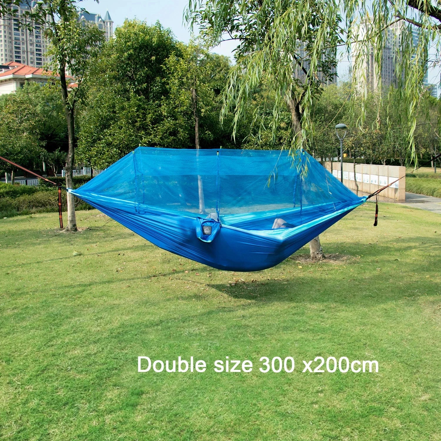 Portable double nylon camping hammock with mosquito net and tree straps set up between trees in a shaded grassy area under a blue sky.