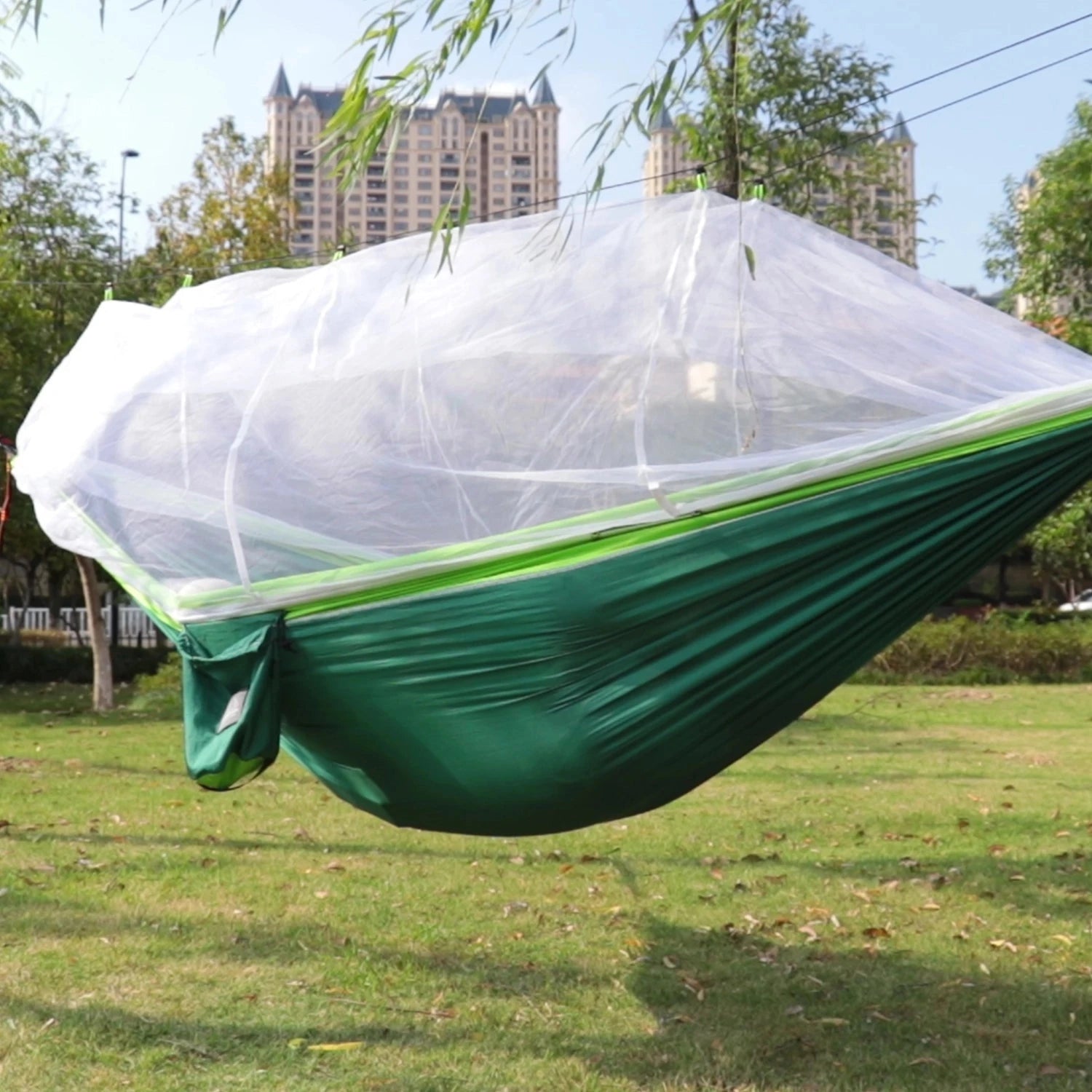 Portable double nylon camping hammock with mosquito net and tree straps set up between trees in a grassy area with a clear sky and shade, surrounded by lush greenery.