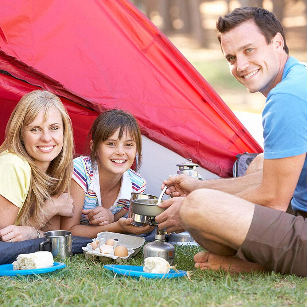 A family enjoying a summer camping trip, gathered around a portable gas stove on the grass, smiling and interacting happily.