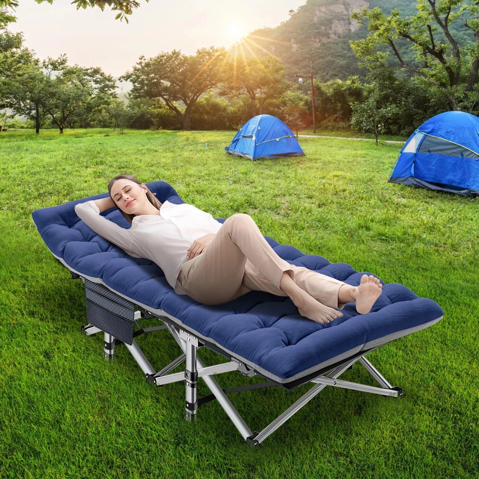 Portable lightweight folding camping cot set up outdoors on grass, near trees and a tent, with a yellow shade in the background under a blue sky.