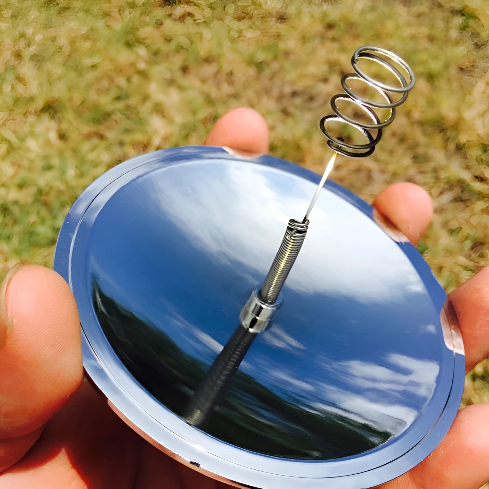 A hand holding a compact concave mirror igniter under sunlight with grass in the background.