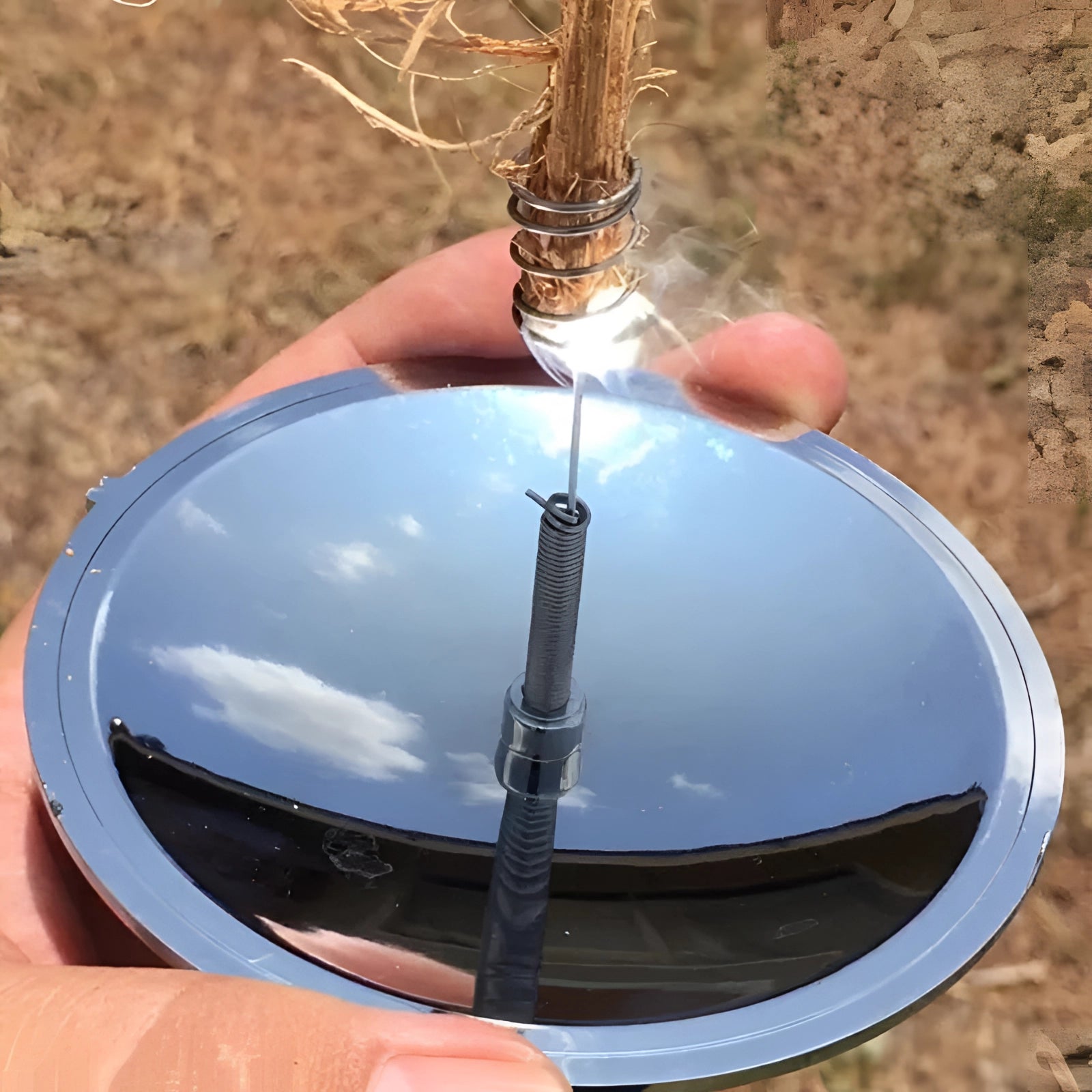 Portable solar fire starter with compact concave mirror igniter placed on grass, surrounded by trees and plants.