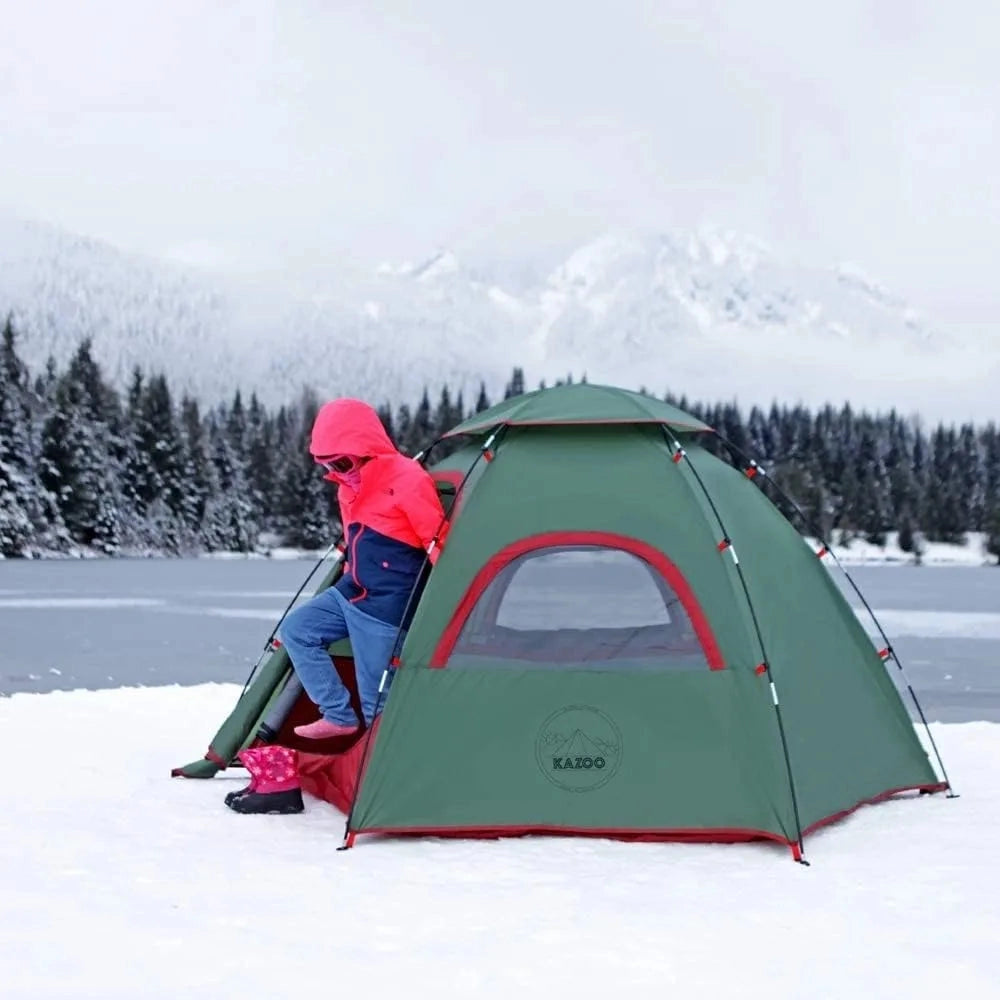 A waterproof family camping tent with dual entrances set up on a snowy mountain slope, surrounded by trees under a clear sky.