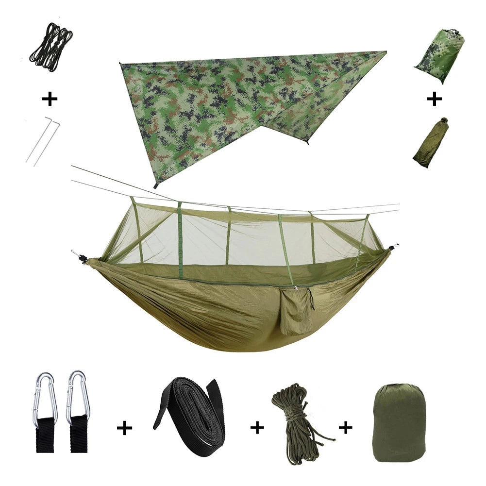 Army green waterproof camping hammock with mosquito net and rain fly, set up outdoors between trees.