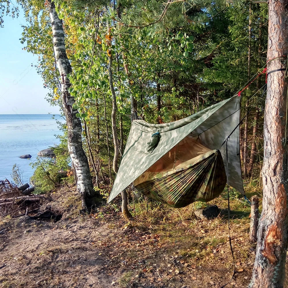 Waterproof camping hammock with mosquito net and rain fly set up in a lush, green forest with tall trees and abundant vegetation, providing shade and a scenic natural landscape.