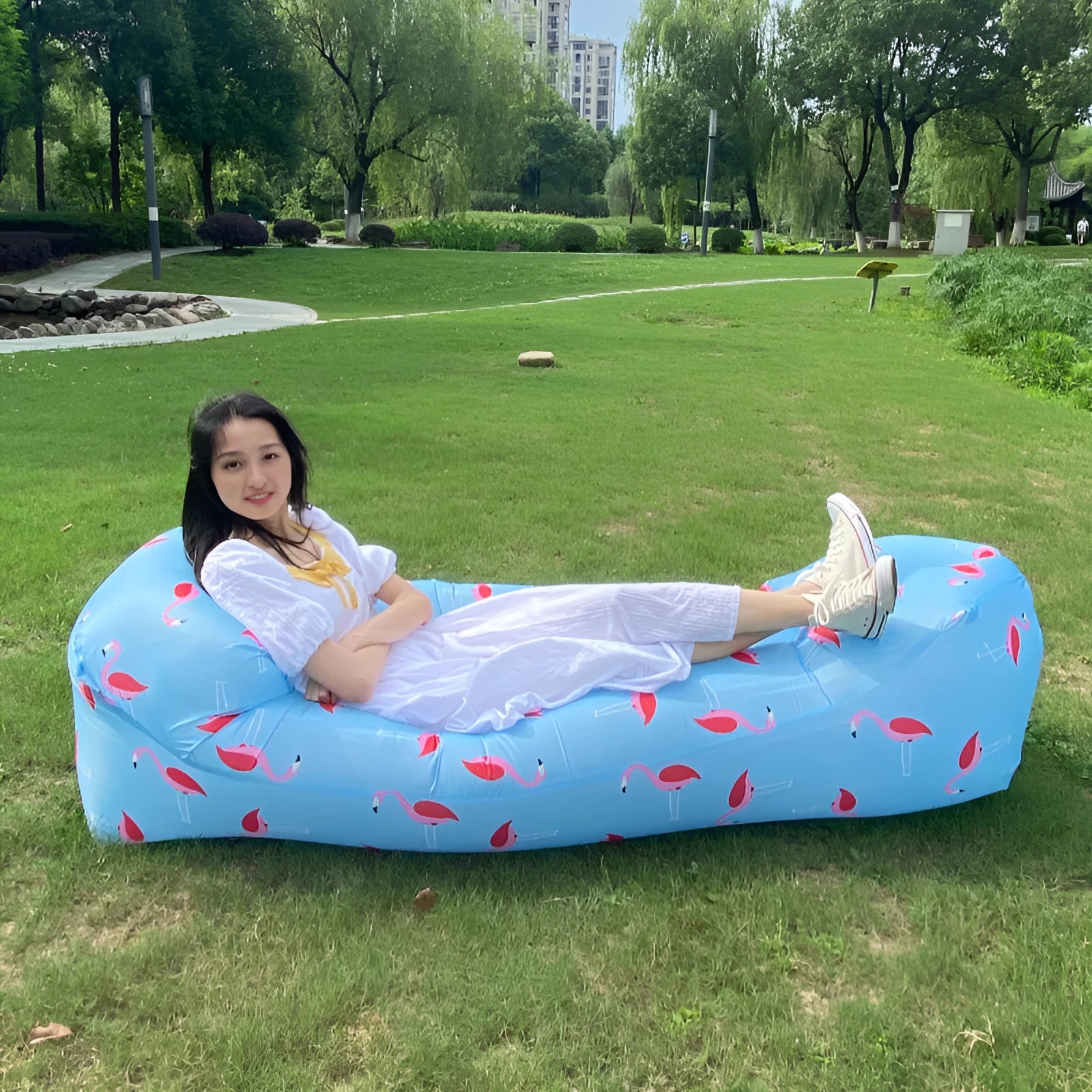 Waterproof outdoor inflatable sofa in a green grassy area with trees and plants in the background, people sitting and enjoying leisure time, a building visible in the distance.