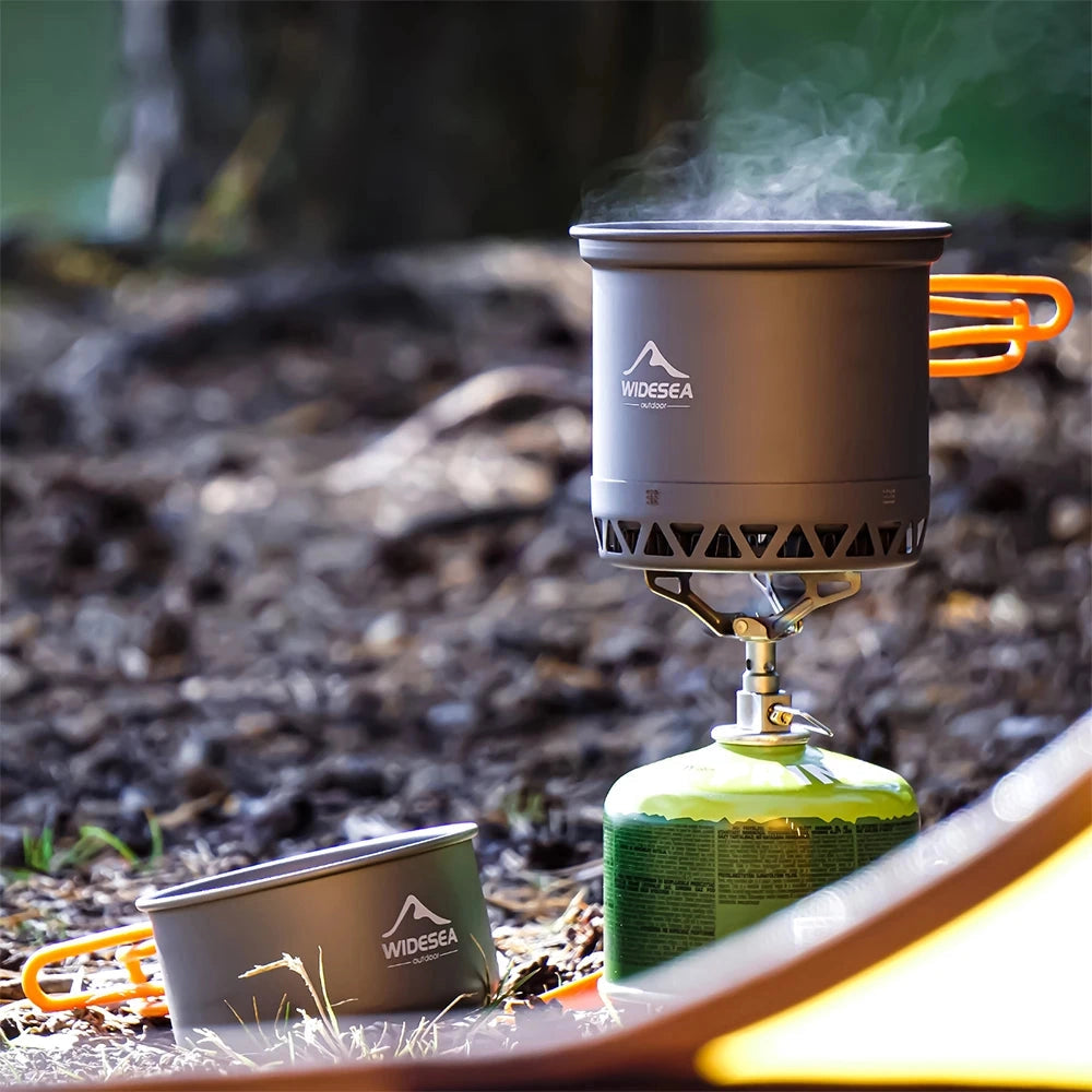 A compact camping stove and portable cookware set placed on a grassy surface, including a gas cylinder and various drinkware items alongside it.