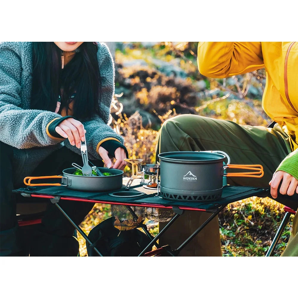 Portable camping table set up on grass with various camping and cooking accessories placed on top, surrounded by outdoor landscape. The table is compact, lightweight, and easily foldable, ideal for recreational outdoor activities.